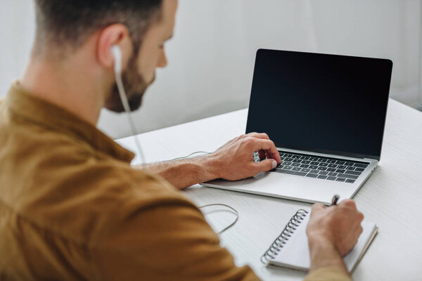 selective focus of man listening music, writing in notebook and using laptop 