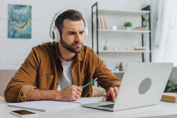 good-looking man listening music, holding pencil and using laptop 