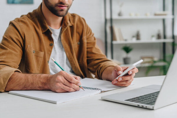 cropped view of man using smartphone and writing in notebook 