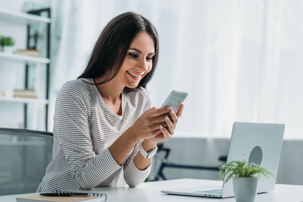 attractive and brunette woman smiling and using smartphone in apartment 