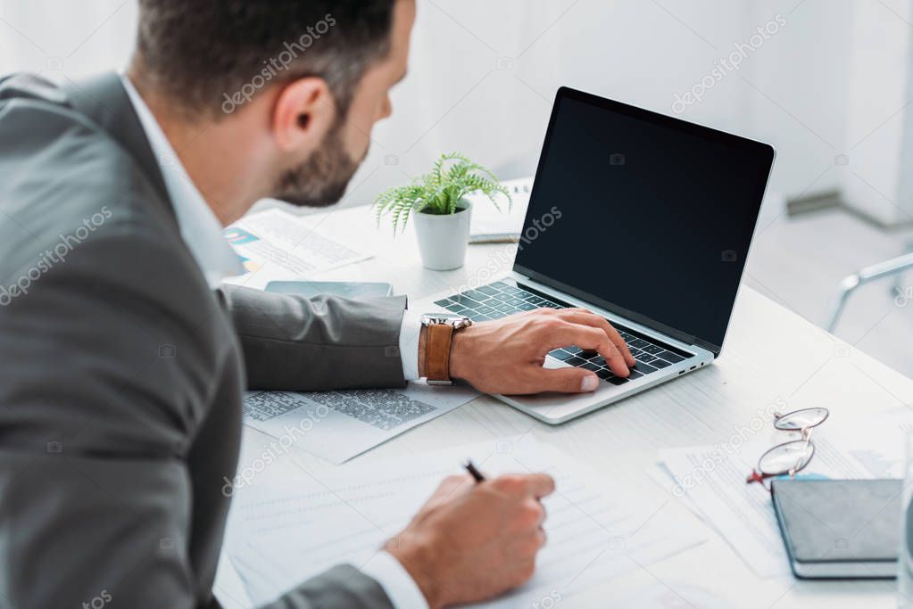 selective focus of man in formal wear holding pen and looking at screen of laptop 
