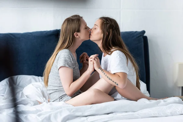 side view of two lesbians holding hands and kissing on bed