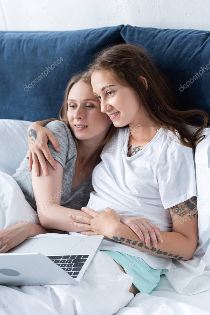 two smiling lesbians embracing while using laptop in bed in morning