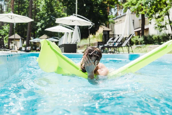 bearded man swimming in swimming pool with green inflatable ring and covering face with hands