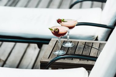 selective focus of cocktail glasses with drinks on wooden table near deck chairs  clipart