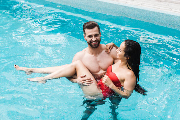 happy bearded man holding in arms brunette woman in red swimwear 