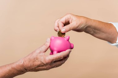 cropped view of retired man holding pink piggy bank while senior wife putting coin isolated on beige  clipart