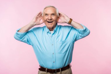cheerful senior man touching ears and smiling while standing on pink  clipart