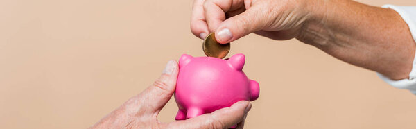 panoramic shot of retired man holding pink piggy bank while senior wife putting coin isolated on beige 