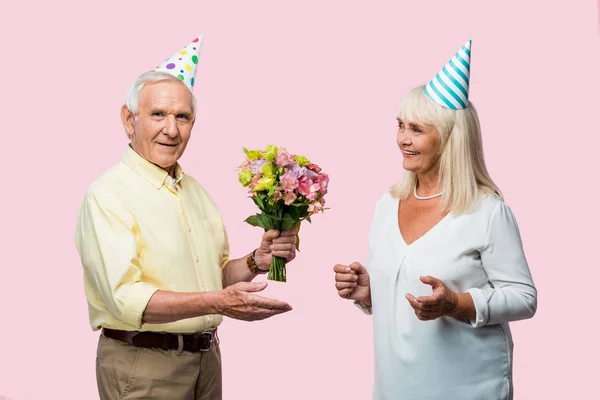 happy senior man in party cap holding flowers near cheerful wife isolated on pink