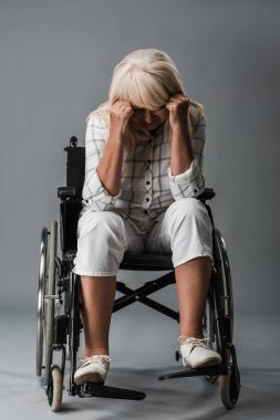 upset retired woman sitting in wheelchair and touching head on grey clipart