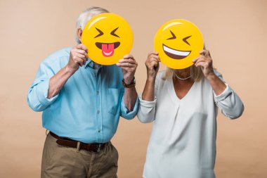 KYIV, UKRAINE - JUNE 14, 2019: retired couple covering faces with yellow happy smileys isolated on beige  clipart