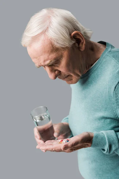 心烦意乱的退休男子看着药片 而拿着玻璃水隔离在灰色 — 图库照片