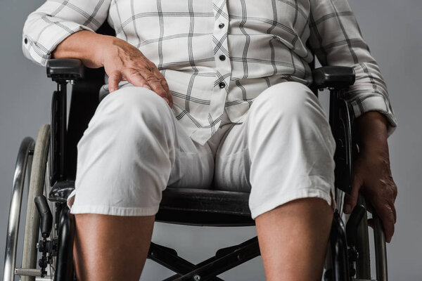 cropped view of disabled retired woman sitting in wheelchair and touching knee isolated on grey 
