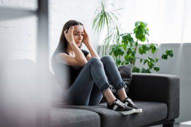 selective focus of young woman suffering from depression while sitting on couch and holding hands near head clipart