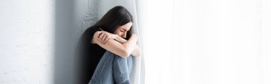 panoramic shot of depressed young woman crying while sitting by window and hiding face in crossed arms clipart