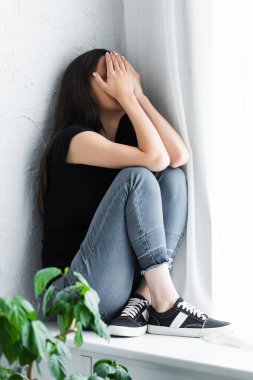 depressed young woman crying while sitting on window sill and covering face with hands clipart