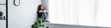 çapraz kolları ile pencere eşik üzerinde oturan ve uzağa bakarak depresif genç kadının panoramik çekim 