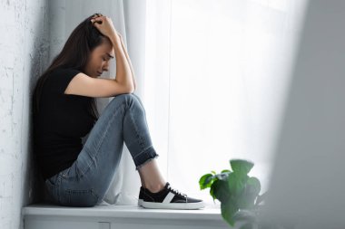 selective focus of young woman crying while sitting on window sill and suffering from depression clipart