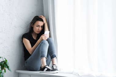 depresif genç kadın evde pencere eşik üzerinde oturan ve akıllı telefon kullanarak