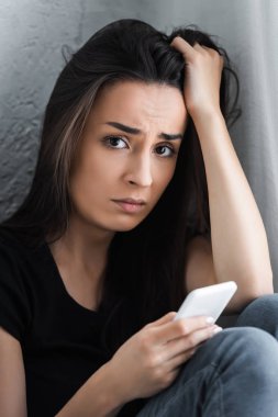depresif genç kadın baş el tutarak ve kamera bakarken akıllı telefon kullanarak
