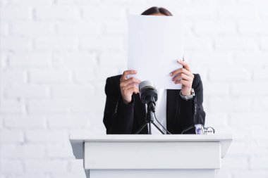 scared lecturer suffering from logophobia and hiding face behind paper while standing at podium tribune clipart