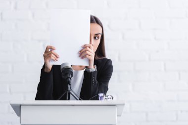 young lecturer suffering from fear of public speaking hiding face with paper sheet while standing on podium tribune clipart