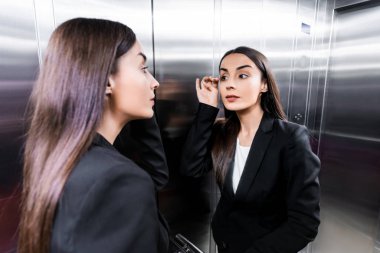 attractive businesswoman looking at reflection in elevator and touching eyelashes clipart