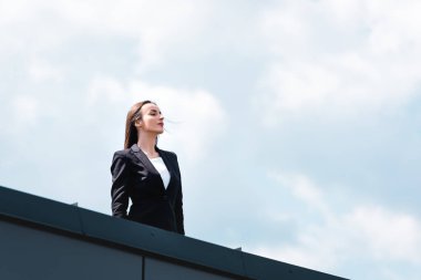 attractive, confident businesswoman smiling and looking away while standing on rooftop clipart