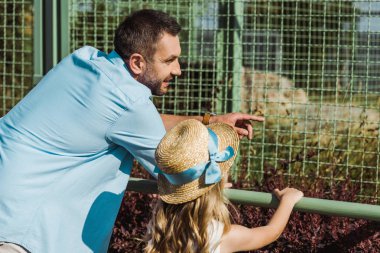 handsome man pointing with finger at cage while standing with daughter in zoo  clipart