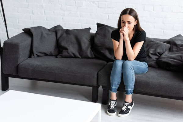 красивая молодая женщина страдает от депрессии, сидя дома на диване и держа руки возле лица
