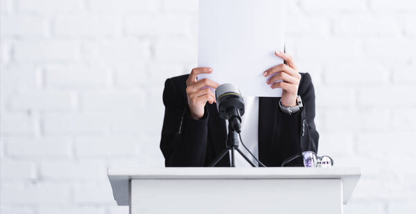 young lecturer, suffering from fear of public speaking, standing on podium tribune and hiding face behind paper sheet