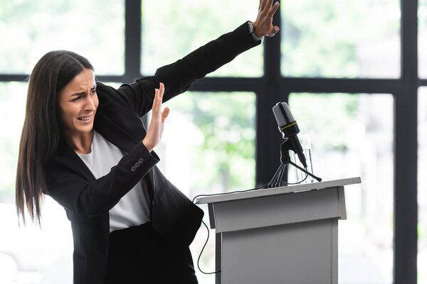 scared lecturer suffering from fear of public speaking gesturing with hands while standing on podium tribune in conference hall