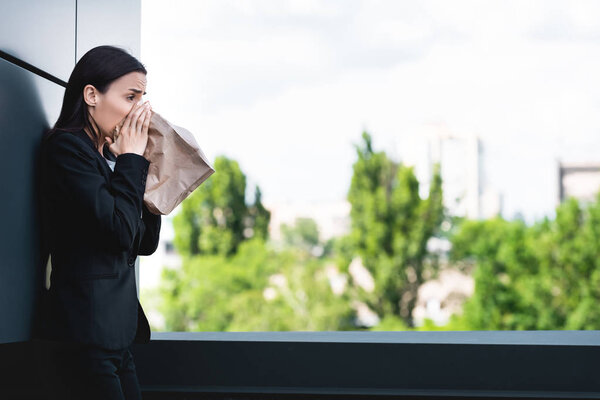 young businesswoman standing on rooftop and breathing into paper bag while suffering from panic attack