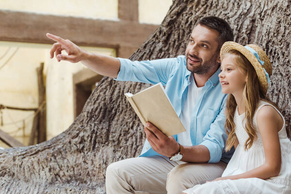 cheerful father holding book and pointing with finger near cute daughter in straw hat 