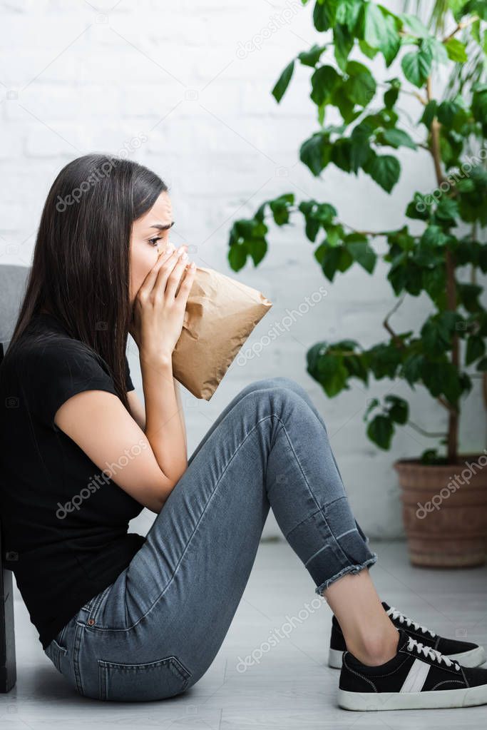 side view of young woman sitting on floor and breathing into paper bag while suffering from panic attack