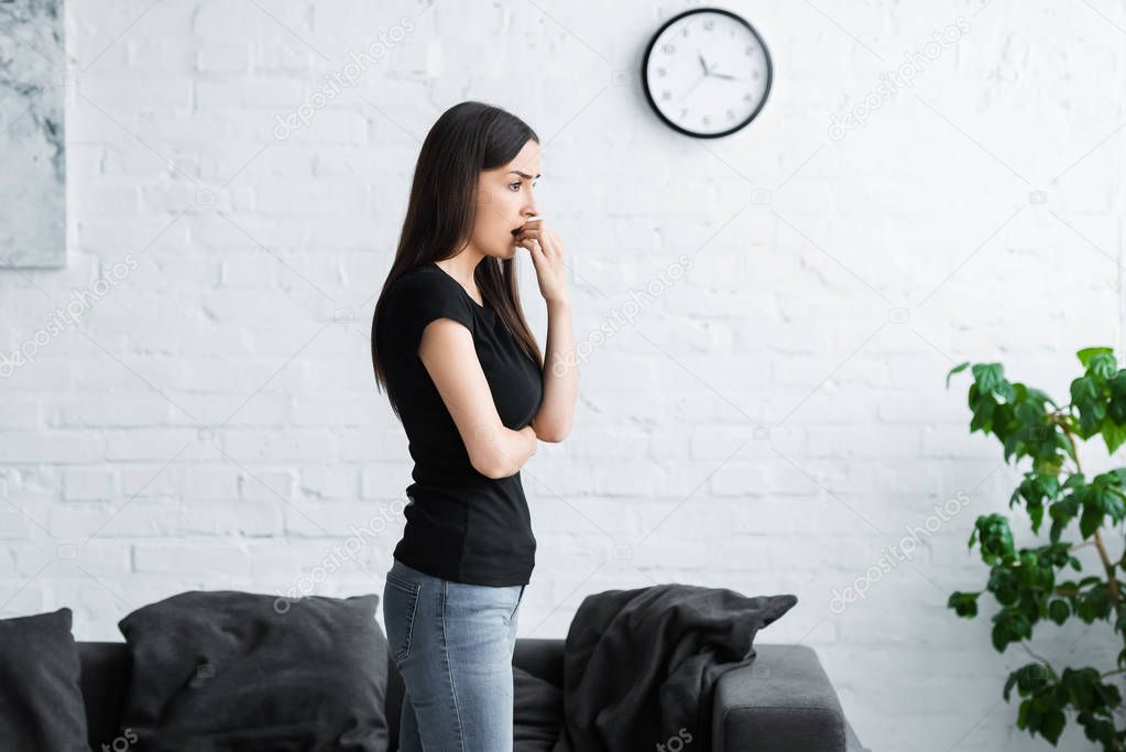 worried young woman looking away while standing at home and holding hand near face