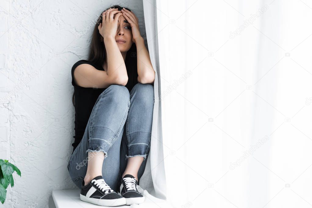 scared young woman suffering from panic attack while sitting on window sill and covering face with hands