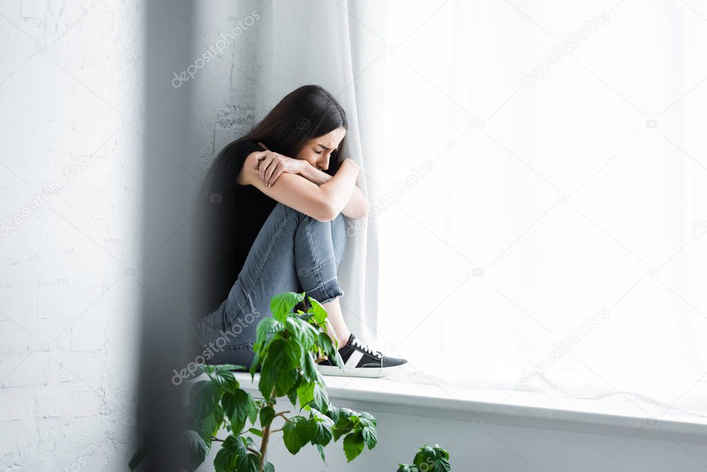 depressed young woman crying while sitting on window sill and hiding face in crossed arms