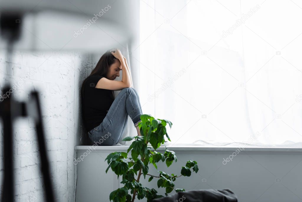 selective focus of depressed young woman crying while sitting on window sill and holding hands on head