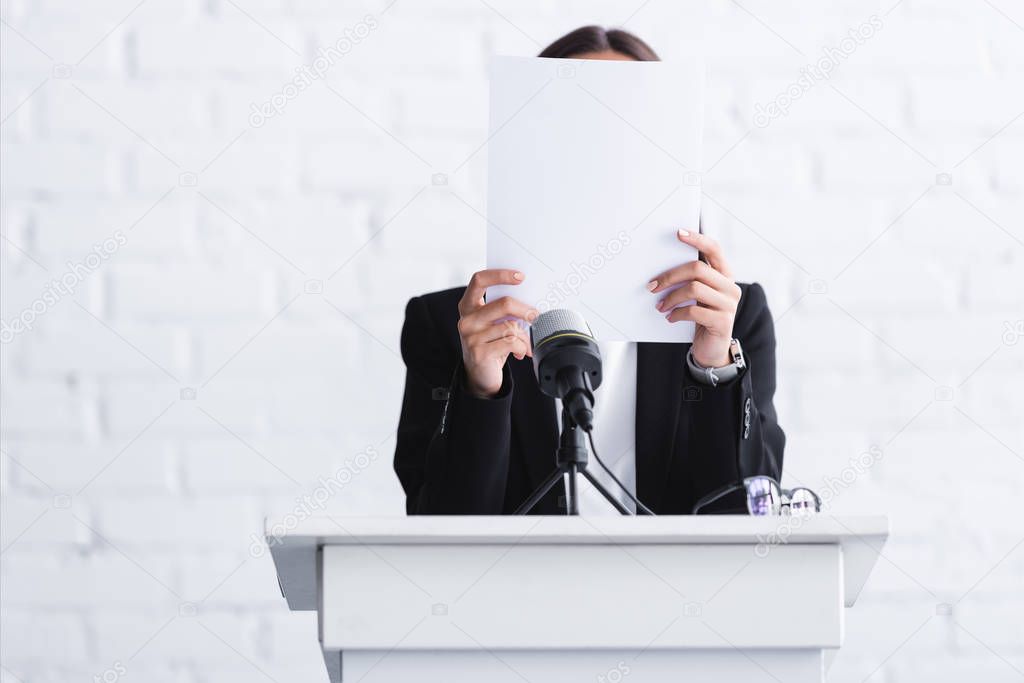 scared lecturer suffering from logophobia and hiding face behind paper while standing at podium tribune