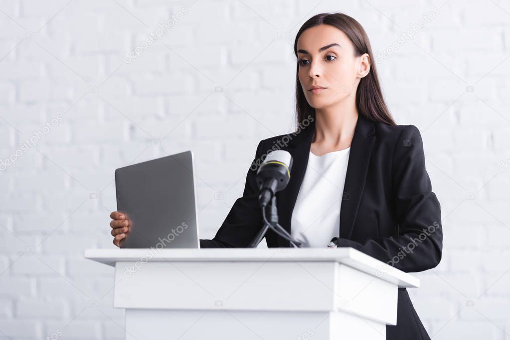 attractive, confident lecturer standing on podium tribune near microphone and laptop