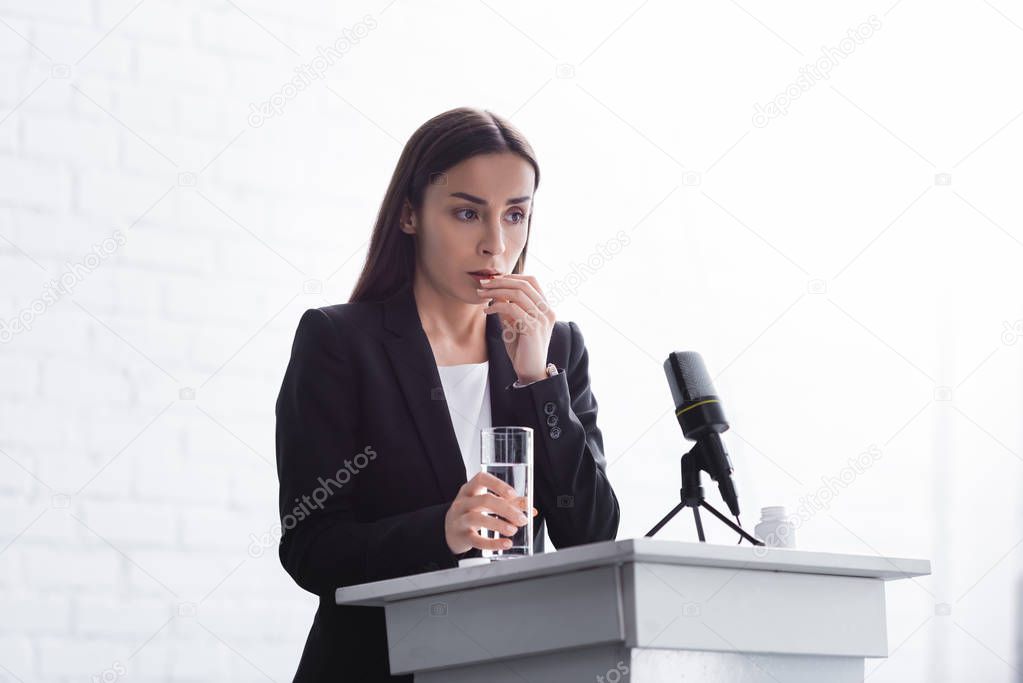 young lecturer suffering from speech anxiety standing on podium tribune with glass of water