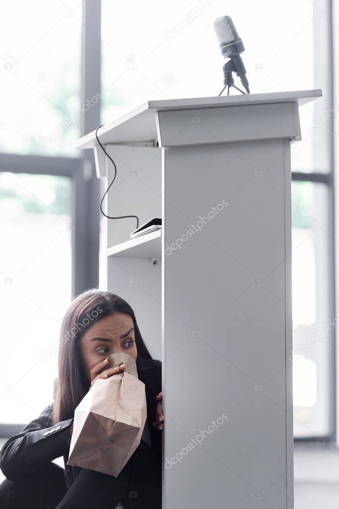 frightened lecturer breathing into paper bag while sitting on floor in conference hall and suffering from panic attack