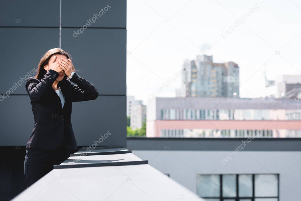 young businesswoman, suffering from fear of heights, standing on rooftop and covering face with hands