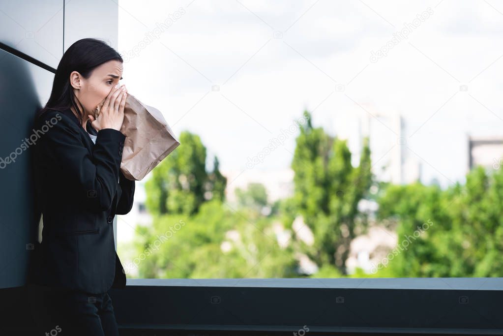 young businesswoman standing on rooftop and breathing into paper bag while suffering from panic attack