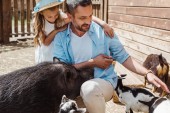 fröhlicher Mann berührt Ziege neben süßer Tochter und Wildschwein im Zoo 