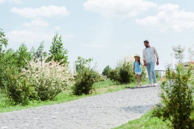 handsome father and daughter walking near green plants and holding hands  clipart