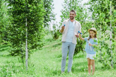 selective focus of cheerful father and daughter holding bubble wands while standing near trees  clipart