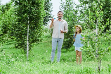 selective focus of father and daughter blowing soap bubbles near trees clipart